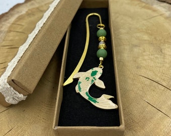 May include: A gold-toned metal bookmark featuring a koi fish charm with cream and green enamel details. The top of the bookmark is adorned with two matte green beads and a small central bead, separated by gold-toned metal spacers, all resting in a box.
