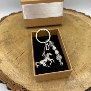 May include: A silver-toned metal keyring featuring a horse charm, a letter D charm, and a strand of white and clear crackle glass beads. The keyring is presented inside a small, rectangular cardboard gift box with a black velvet insert.