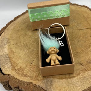 Könnte beinhalten: Ein kleiner, blauhaariger Troll-Schlüsselanhänger mit einem silbernen Anhänger, auf dem "B" steht, in einer braunen Geschenkbox mit grünem Band.