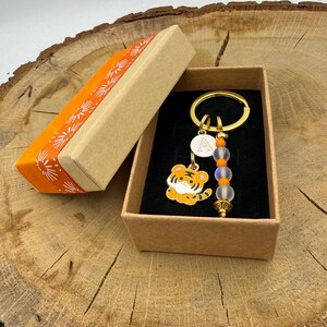 Op de afbeelding: Een goudkleurige metalen sleutelhanger met een kleine oranje en witte tijgerbedel, een witte ronde bedel met de letter A en een streng van doorschijnende en oranje kralen, gepresenteerd in een rechthoekige geschenkdoos met een oranje deksel.
