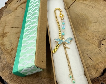 May include: A gold-toned metal bookmark featuring a dragonfly charm with light blue enamel wings, iridescent beads, and a small clear crystal accent. The bookmark is presented in a rectangular cardboard gift box with a green patterned ribbon.