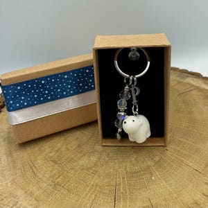 Op de afbeelding: Een witte ijsbeer sleutelhanger met een zilveren sleutelring en kralenaccenten, gepresenteerd in een kleine bruine geschenkdoos met een blauw en wit stippenlint. De doos is open en toont de sleutelhanger.
