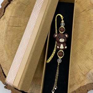 Op de afbeelding: Een bruine teddybeer bladwijzer met een goudkleurig metalen ontwerp, gepresenteerd in een bruine geschenkdoos met een patroonlint. De bladwijzer heeft een kleine berenfiguur en een bungelende ketting.