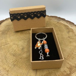 Puede incluir: Una caja de cartón marrón con un borde de encaje negro contiene un llavero plateado con un dije de orangután naranja, un dije de letra "B" plateado y cuentas naranja y transparentes.
