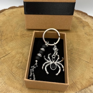 Könnte beinhalten: Ein silberner Spinnen-Schlüsselanhänger mit einem schwarzen Edelstein-Mittelstück und einem Perlenakzent, präsentiert in einer kleinen Geschenkbox mit schwarzem Band. Der Schlüsselanhänger wird auf einer Holzoberfläche präsentiert.