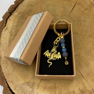 Op de afbeelding: Een goudkleurige draak sleutelhanger met een gouden ring, blauwe kralen en een kleine gouden bedel. De sleutelhanger wordt gepresenteerd in een bruine geschenkdoos met een zilveren lint. De doos staat op een houten oppervlak.