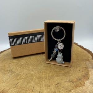 Puede incluir: Un llavero de metal plateado con un pequeño dije de gato gris, un dije circular blanco con la letra N y una hilera de cuentas grises e iridiscentes. El llavero se presenta dentro de una pequeña caja de regalo rectangular de cartón marrón.