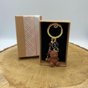 Pode incluir: Um porta-chaves dourado com um pendente de urso castanho texturado, um pequeno pendente circular preto com a letra Y em branco e várias contas castanhas e metálicas, apresentado numa caixa de presente retangular de cartão com fita branca.