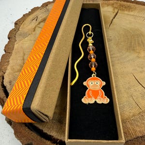 May include: A gold-toned metal bookmark featuring a dangling charm of an orange monkey. The charm is accented with three clear faceted beads and small orange spherical beads. The bookmark is presented in a long, narrow cardboard gift box.