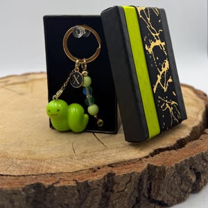 May include: A gold-toned keyring featuring a bright green caterpillar charm, a small circular black charm with the letter X, and a strand of light green and iridescent beads, presented in a black gift box with a lime green and black patterned ribbon.