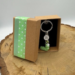 Op de afbeelding: Een groene rups sleutelhanger met een zilveren sleutelring en een letter "B" bedel, gepresenteerd in een kleine bruine geschenkdoos. De doos is versierd met een lichtgroen lint met een wit bloemmotief. Achtergrond is hout.