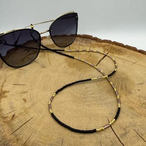 Op de afbeelding: Een cat-eye zonnebril met donkere glazen en goudkleurige monturen, bevestigd aan een kralenketting. De ketting heeft een herhalend patroon van kleine zwarte kraaltjes, goudkleurige tussenkralen en langwerpige koperkleurige buisjes.
