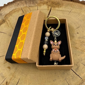 May include: A gold-toned keyring featuring a brown kangaroo figurine with a joey, a small black circular charm with the letter E, and a strand of three beads in beige and translucent grey, presented in a rectangular cardboard gift box.