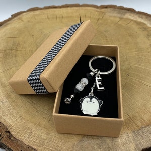 Op de afbeelding: Een bruine doos met een zwart-wit geruit patroon bevat een zilveren pinguïn sleutelhanger met een zilveren letter E bedel en heldere kristallen kralen.