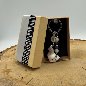 May include: A keychain featuring a small, sculpted pigeon figurine with grey and white feathers, a silver-toned metal ring, a circular charm with the letter A, and three spherical beads in shades of grey and white, presented in a tan gift box.