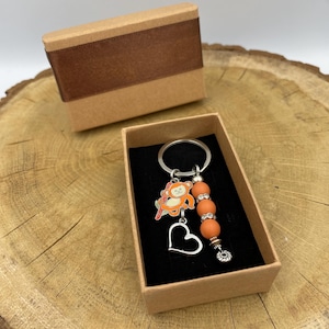 May include: A keychain featuring a monkey charm, orange beads, and a silver heart pendant, displayed in a small gift box with a brown ribbon. The keychain is on a silver ring. The box is brown.