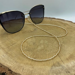 Op de afbeelding: Zwarte cat-eye zonnebril met gouden details en een gouden kralenketting. De zonnebril heeft verloopglazen. De ketting is gemaakt van kleine, goudkleurige kralen. De zonnebril en ketting liggen op een houten oppervlak.