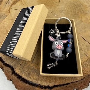 Op de afbeelding: Een grijze ezel sleutelhanger met een zilveren sleutelring en een kralen accent, gepresenteerd in een bruine geschenkdoos met een zwart-wit gestreept lint. De ezel heeft roze oren en een lachend gezicht.