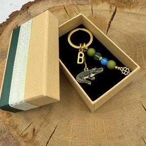 Op de afbeelding: Een goudkleurige metalen sleutelhanger met een letter B-bedel, een groene alligatorbedel en een streng van drie kralen in matgroen en iriserend blauw, eindigend in een klavertjevier-bedel, gepresenteerd in een beige kartonnen geschenkdoos.