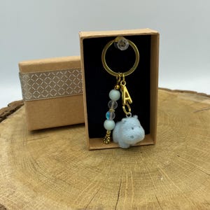 Può includere: Un portachiavi color oro con un ciondolo a forma di ippopotamo azzurro, una lettera "A" dorata e una fila di perline azzurre e iridescenti. Il portachiavi è presentato in una piccola scatola regalo marrone con un nastro decorativo.