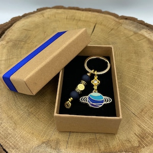 Puede incluir: Un llavero de planeta azul y dorado con un dije de cuentas negras y doradas. El llavero está en una caja de cartón marrón con una cinta azul.