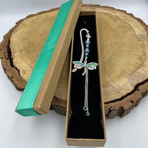 Può includere: Un segnalibro a forma di libellula argentata con ali smaltate colorate e una catena pendente, presentato in una confezione regalo marrone con un nastro verde acqua. Il segnalibro presenta un accento di perline.