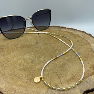 Op de afbeelding: Witte en lichtpaarse kralenketting voor brillen met gouden kralen en een gouden bedel. De ketting ligt op een houten oppervlak met een zwarte zonnebril.
