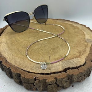 Op de afbeelding: Een witte, roze en groene kralenketting voor een bril met een zilveren bedel. De ketting ligt op een houten oppervlak met een zwarte zonnebril op de achtergrond.
