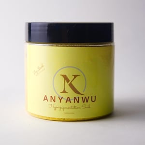 May include: A jar of Anyanwu Hyperpigmentation Scrub with a yellow label and a black lid. The label features a stylized "K" inside a circle, with the brand name and product description in brown text. The jar is filled with a yellow scrub.