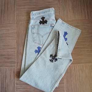 Op de afbeelding: Lichtblauwe spijkerbroek met zwarte en blauwe kruisvormige patches. De jeans heeft een klassiek ontwerp met vijf zakken en een vervaagde wassing. Kruispatches zitten op de achterzak en de voorkant van de benen.