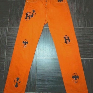 Könnte beinhalten: Orangefarbene Jeans aus Denim mit schwarzen, kreuzförmigen Patches. Die Jeans haben eine gerade Beinweite und einen Knopfverschluss.