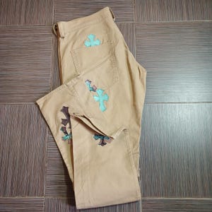 Op de afbeelding: Beige broek met een decoratief kruisontwerp aan de voor- en achterkant. Het kruis is lichtblauw.