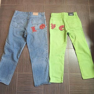 May include: Two pairs of jeans. One pair is light blue denim with red patches on the back pockets. The other pair is bright green with red patches on the back pockets. Both pairs have the Levi's brand tag.