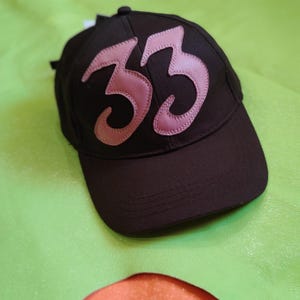 Peut inclure: Casquette de baseball noire avec le chiffre "33" en rouge-rosé. Le chiffre est cousu sur la casquette. La casquette est posée sur une surface verte.