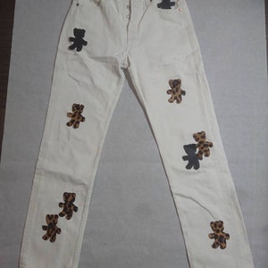 May include: Off-white denim jeans with a button fly and multiple bear patches. Some bear patches are solid black, while others feature a brown and tan leopard print pattern. The jeans have a classic five-pocket design.
