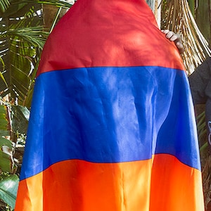 May include: A person holding a red, blue, and orange flag. The flag is draped over the person's shoulders and is made of a soft, flowing fabric.