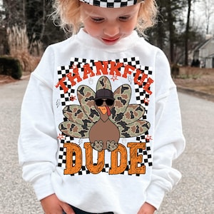 Pode incluir: Camisola branca com um desenho de um peru usando óculos de sol e um chapéu. As penas do peru têm um padrão de camuflagem. As palavras "THANKFUL DUDE" são impressas em laranja e preto.