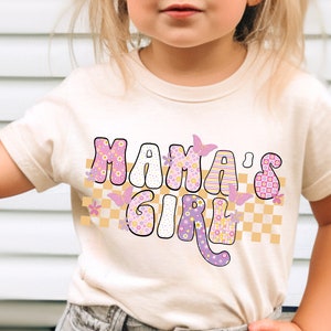Op de afbeelding: Een licht beige t-shirt met een roze en gele afbeelding met de tekst "Mama's Girl" met vlinders en een geruit patroon.