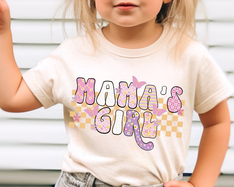 Op de afbeelding: Een licht beige t-shirt met een roze en gele afbeelding met de tekst "Mama's Girl" met vlinders en een geruit patroon.