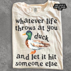 May include: An ivory t-shirt featuring a watercolour illustration of a mallard duck with a green head and brown body. The black text reads: whatever life throws at you duck and let it hit someone else. A label at the neck reads Comfort Colors.