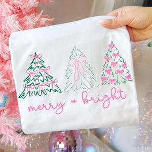 Könnte beinhalten: Weißes T-Shirt mit einem festlichen Design mit drei grünen und rosa Weihnachtsbäumen. Die Bäume sind mit rosa Schleifen verziert, und der Schriftzug "merry & bright" steht darunter in Rosa.