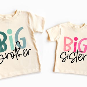 May include: Two cream colored t-shirts with the words "Big Brother" and "Big Sister" printed on them in black and colorful lettering.