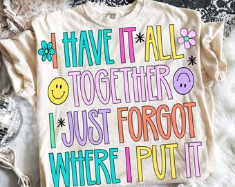 Può includere: T-shirt color crema con la scritta colorata e giocosa "I HAVE IT ALL TOGETHER I JUST FORGOT WHERE I PUT IT". Il design include faccine sorridenti e grafiche floreali. Una maglietta divertente e casual.