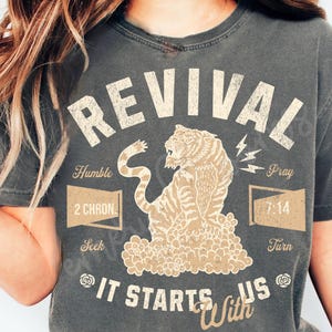 May include: A gray t-shirt with a vintage-style graphic design featuring a tiger, the word "REVIVAL", and the text "It Starts With Us". The design also includes the words "Humble", "Seek", "Pray", "Turn", and "2 Chron" and "7:14".