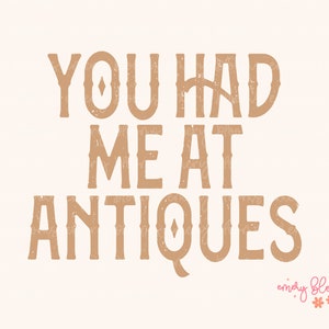 May include: A beige print with the words "YOU HAD ME AT ANTIQUES" in a brown, textured font. The bottom right corner features the text "emery blooms" in a script font with floral accents.