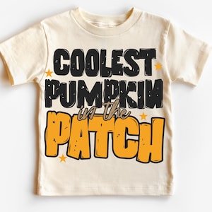Può includere: Una maglietta color crema con la scritta "COOLEST PUMPKIN in the PATCH" in nero e arancione. Il testo è delineato in nero e arancione, con piccole stelle gialle. La maglietta è a maniche corte e girocollo.