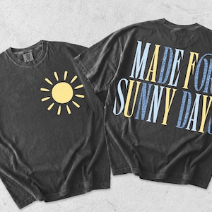 May include: Two charcoal grey t-shirts. One has a yellow sun graphic on the front. The other has the text "MADE FOR SUNNY DAYS" in yellow and blue on the back. The shirts are made of a soft material.
