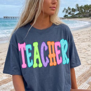 Peut inclure: Un t-shirt bleu marine avec le mot "TEACHER" en grosses lettres colorées à bulles. Les lettres sont bordées de rose, de vert, d'orange et de bleu, avec un effet pailleté. Le t-shirt est porté par une personne sur une plage.