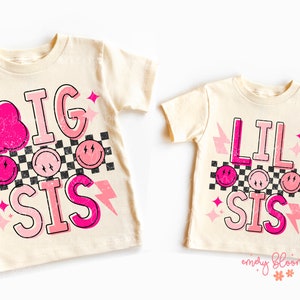 May include: Two cream colored t-shirts with pink and black graphic designs. The larger shirt says "BIG SIS" and the smaller shirt says "LIL SIS". Both shirts feature a checkered pattern, lightning bolts, and smiley faces.
