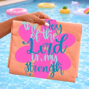 May include: A folded peach-colored t-shirt with a large pink flower design. The flower contains the text "the Joy of the Lord is my Strength" in turquoise and blue script. The shirt is held up against a pool background.